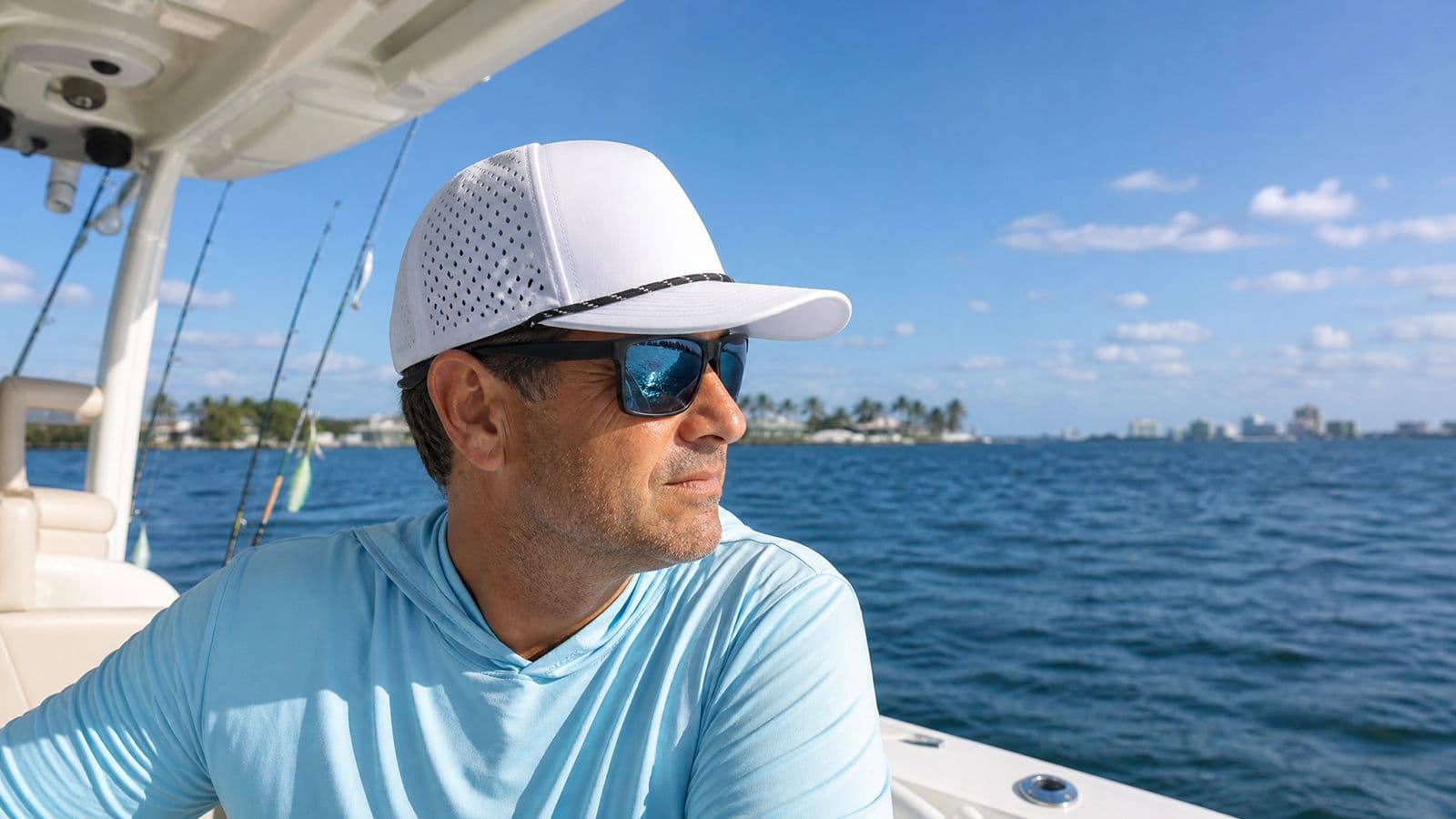 A man in sunglasses looks out over the water from a boat, wearing a white rope hat with a dark cord trim along the bill and perforated side panels.