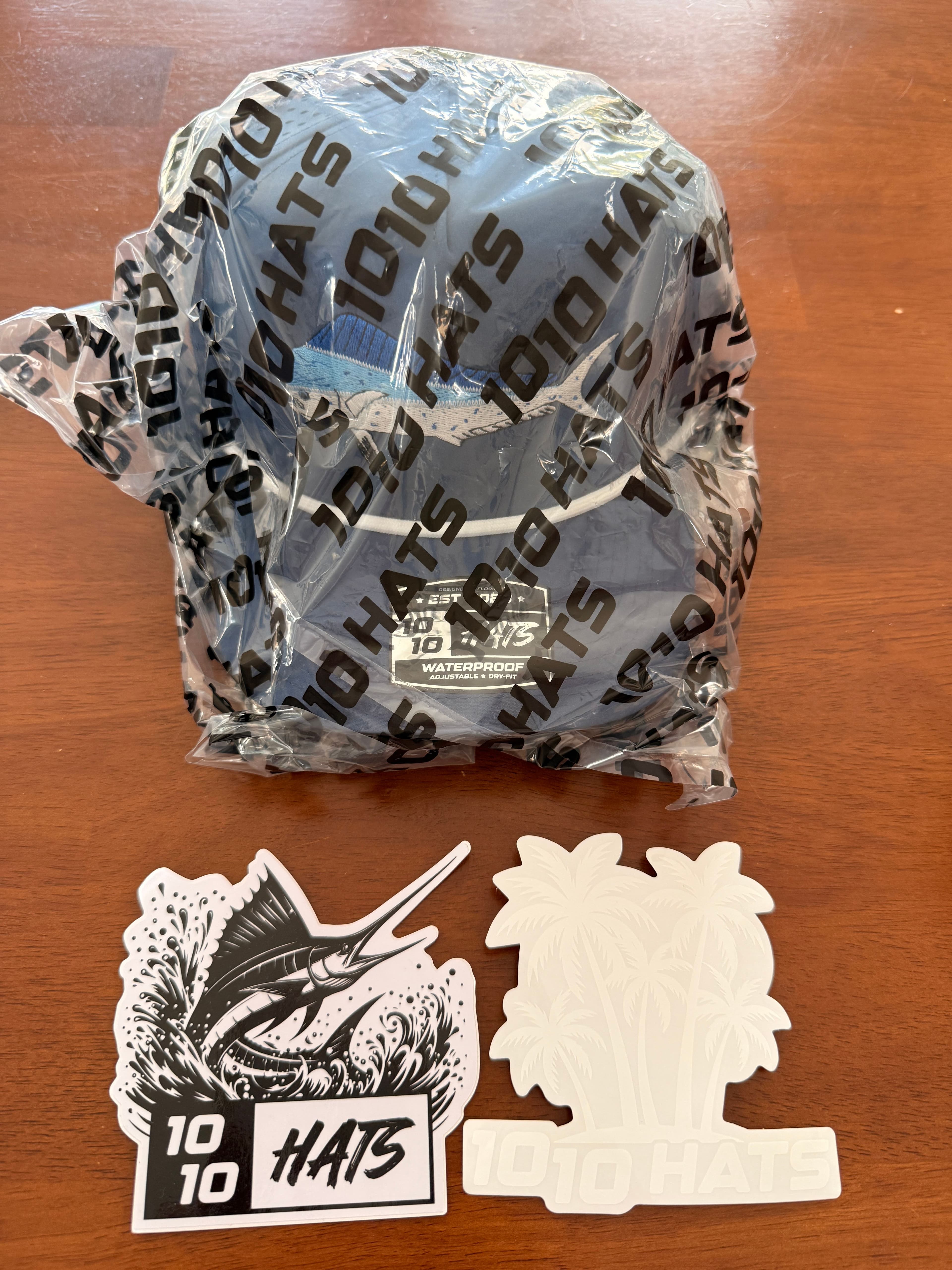 The wrapped 10-10 Hats Mahi Fish Hat next to its two included stickers — a palm-tree wordmark decal and a swordfish-and-wave decal